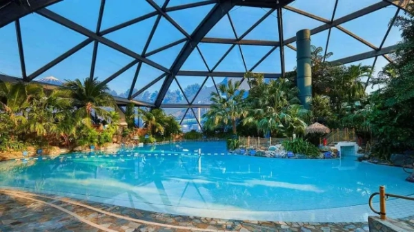 Басейн Center Parcs у селищі Шервудський ліс з великою скляною куполоподібною стелею та тропічними деревами навколо води.