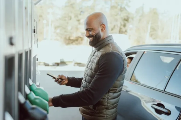 Усміхнений зрілий чоловік з поголеною головою здійснює онлайн-платежі, стоячи біля машини на заправці