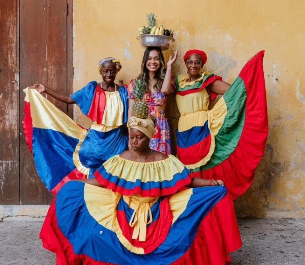 Худа з трьома колумбійськими жінками в барвистих традиційних сукнях. Вона в центрі, тримаючи над головою миску з фруктами.