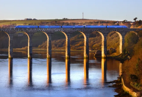 Вид на синій поїзд Lumo, що їде над річкою по високому арочному мосту.