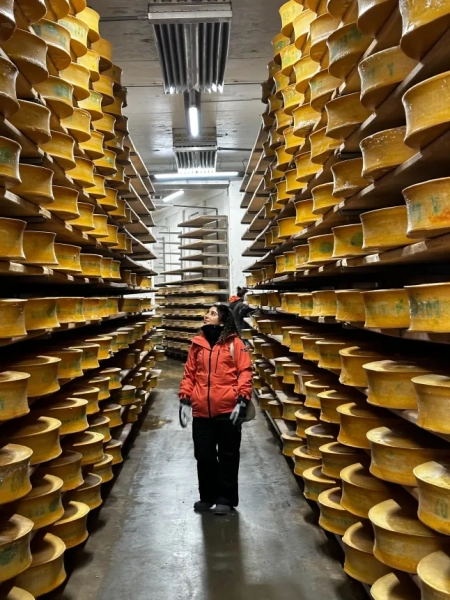 Зінеб Лазрак стоїть у вузькому коридорі з сирних коліс, що тягнуться до стелі. На ній помаранчева лижна куртка.