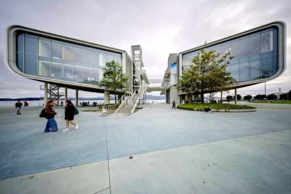 Вражаючий вид на сучасну архітектуру торгового центру Centro Bot??n у Сантандері з його елегантним, футуристичним дизайном, що включає великі скляні панелі та підвісну доріжку, що з'єднує дві секції будівлі. Споруда стоїть на піднятому рівні, спираючись на стрункі колони, а навколо неї ростуть дерева та зелень. Люди гуляють просторою площею, насолоджуючись відкритим простором біля набережної. Хмарне небо додає спокійного та м'якого світла до пейзажу, підкреслюючи інноваційний дизайн будівлі та спокійну атмосферу району. Сантандер, Кантабрія, Іспанія - 18 жовтня 2024 року.