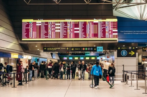 Інтер'єр зони відправлення аеропорту Лісабона, де люди стоять у черзі та ходять під великим табло відправлення з інформацією про рейси.