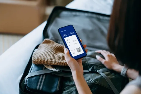Вид через плече молодої азійської жінки, яка користується смартфоном, перевіряє цифровий авіаквиток та посадковий талон на екрані пристрою, пакуючи валізу на ліжку для подорожі. Планування подорожі. Концепція подорожей та відпустки.