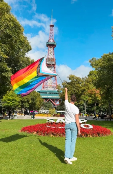 Калум Максвігган розмахує прапором Прайду, стоячи спиною до камери, перед мініатюрною Ейфелевою вежею в Саппоро, Японія.