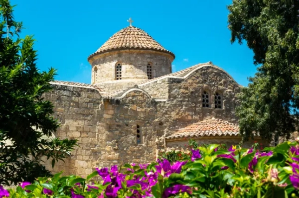 Приголомшливий крупний план церкви Ангелоктісті в Ларнаці, Кіпр, яка має кам'яні стіни та вежу з черепичним дахом і хрестом на вершині. На передньому плані зображені пурпурові квіти та зелень.
