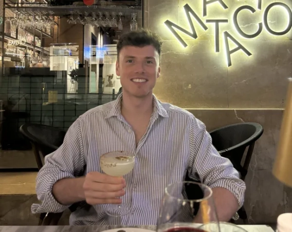 Джеймі Вілсон у сіро-білій смугастій сорочці тримає коктейль у ресторані.