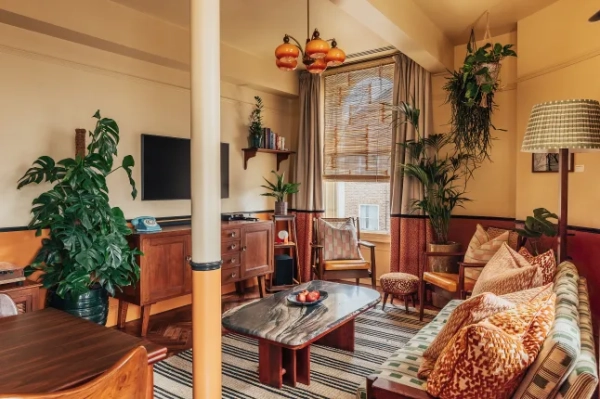 Фотографія інтер'єру житлового простору The Lodgings. Тут багато кімнатних рослин, меблі золотисто-помаранчевого кольору, кам'яний стіл, візерунчастий диван з помаранчевими подушками, книжкова полиця та смугастий килим. Вся кімната дуже тепла, з жовтими стінами.